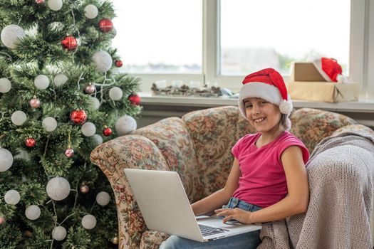 Christmas tree in a large living room. Little girl plays near the Christmas tree. A girl with a laptop communicates with friends.