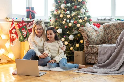 Indoor shot of beautiful happy young woman shopping online on laptop in cozy Christmas interior. Mother on the floor next the Christmas tree and sofa and daughter embrace her, shopping gifts