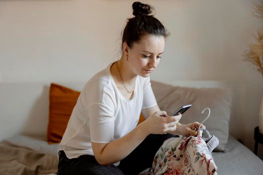 Young woman sitting on the bed at home and taking photo of her old clothes to sell them online. Selling on website, e-commerce. Reuse, second-hand concept. Conscious consumer, sustainable lifestyle