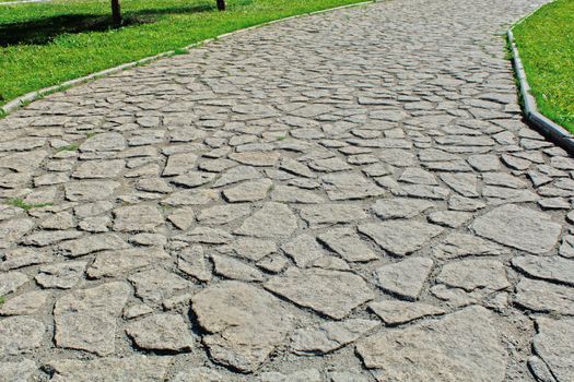The road of gray stones against the background of green lawns in the city park. Place for walking.