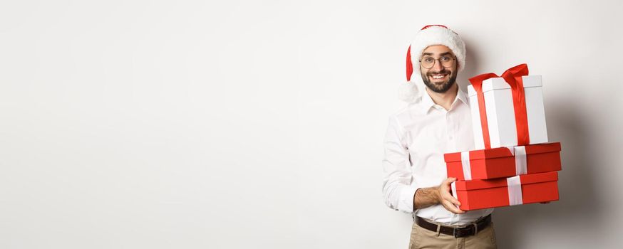 Winter holidays and celebration. Happy guy bring christmas presents, holding gifts and wearing Santa hat, standing over white background.