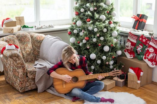 beautiful dark sits near the Christmas tree decorated with balls and light and playing the guitar and singing. Young beautiful smiling woman play guitar