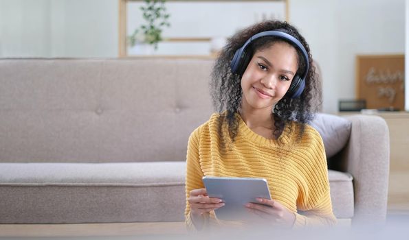 Young satisfied female in casual clothes yellow and headphones surfing digital tablet while sitting on floor with legs crossed near soft couch