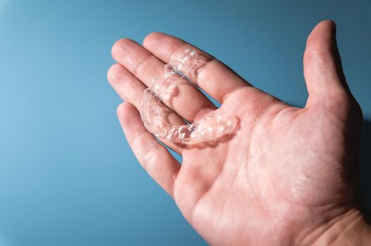 Close-up of a man holding invisible aligners for whitening and straightening his teeth on a blue background. Orthodontic treatment instead of or after braces.