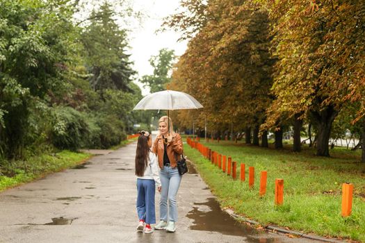 Cheerful mother and her little daughter having fun together in the autumn background under the umbrella. Happy family in the fall background.