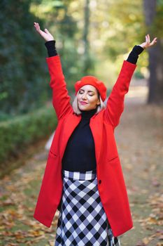 Dreamy female with closed eyes in vivid coat with beret standing with raised arms in park with fallen leaves on autumn day