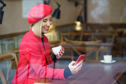 Through glass side vies of smiling female in elegant coat text messaging on cellphone while sitting with cup of coffee at table in cafeteria