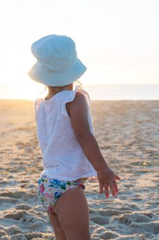 Little girl in a panama hat stands with her back on the beach and looks at the ocean