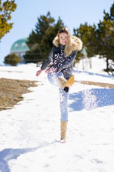 Blonde happy woman kicking snow in a snow-covered forest in the mountains in Sierra Nevada, Granada, Spain. Female wearing winter clothes..