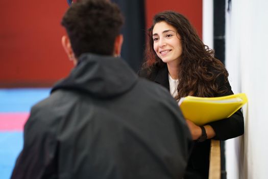 Positive brunette with folder smiling and listening to male athlete during motivational therapy session in gym in daytime