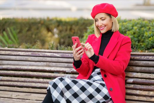 Delighted female in red coat and beret scrolling news on modern cellphone while sitting on wooden bench in autumn park