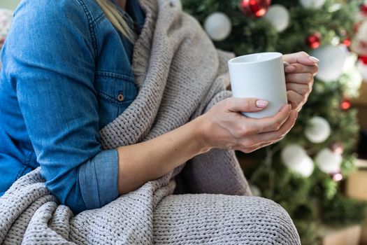 Young woman at home at Christmas time, drinking hot beverage from the mug. Christmas tree in the background. Celebrating Christmas and New Year at home in cozy