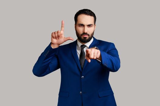 Portrait of strict bearded man pointing at camera and showing lame or loser gesture, mocking your failures, wearing official style suit. Indoor studio shot isolated on gray background.