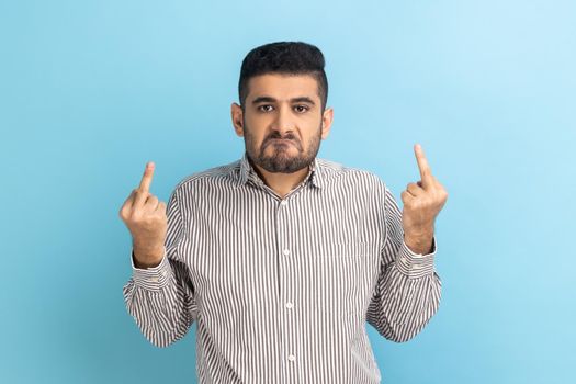 Impolite businessman showing middle fingers and asking to get off looking at camera with negativity, disrespectful behaviour, wearing striped shirt. Indoor studio shot isolated on blue background.