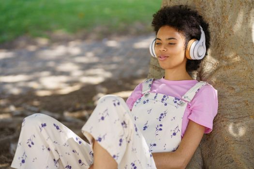 Glad African American female in casual wear listening to song in wireless headphones while sitting near thick tree trunk in park
