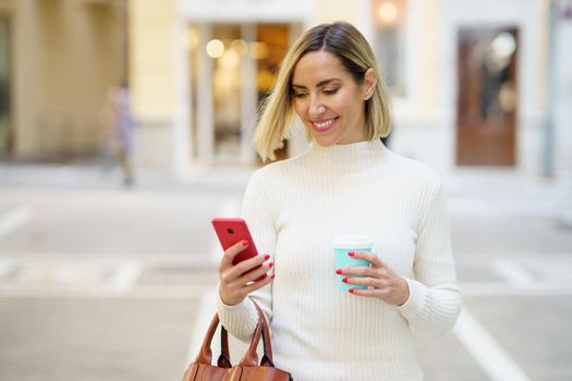 Happy female with blond hair wearing white turtleneck browsing cellphone and holding cup of takeaway beverage