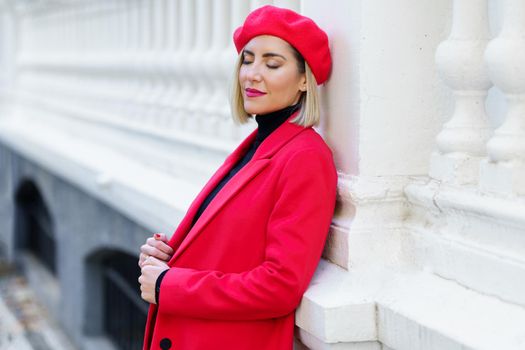 Dreamy female with blond hair in red clothes with beret standing with closed eyes near white fence on street of city