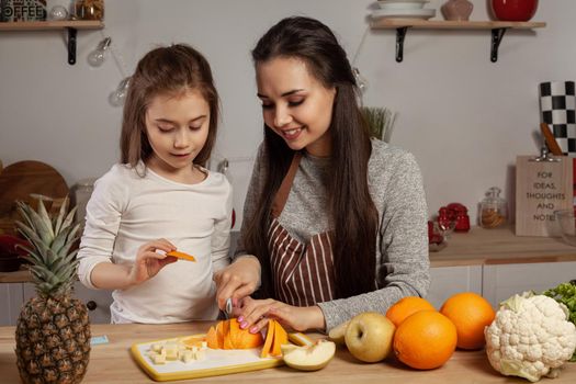 Happy loving family are cooking together. Beautiful mother and her daughter are doing a fruit cutting, smiling and having fun at the kitchen, against a white wall with shelves and bulbs on it. Homemade food and little helper.