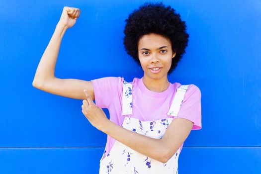 Content muscular African American female in casual wear pointing at bicep and looking at camera against blue background on street
