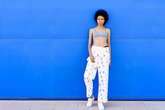 Full body of fit African American female on overall and bra looking away while standing on blue background in city
