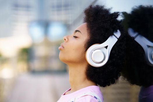 Side view of African American female listening to song in wireless headphones near glass building on street against blurred background