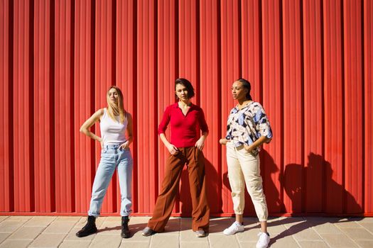 Full body of multiethnic stylish female friends standing with hands in pockets near red wall on sunny street of city