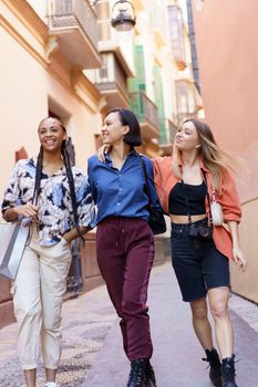 Happy young diverse stylish ladies walking together near residential building after shopping in summer
