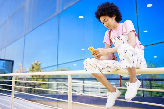 Full body of focused African American female texting message on cellphone while sitting on railing near glass building in city
