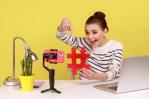Smiling excited woman blogger showing big red hashtag symbol at smartphone camera, sharing viral content, tagged message. Indoor studio studio shot isolated on yellow background.