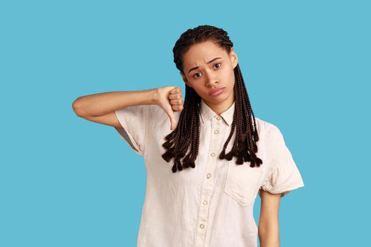 Discontent woman with black dreadlocks showing disapproval sign, keeps thumb down, expresses dislike, frowns face in discontent, wearing white shirt. Indoor studio shot isolated on blue background.