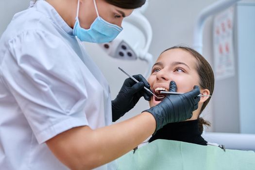 Young teenage female at dental checkup in clinic. Teenage girl sitting in chair, doctor dentist with tools examining patient's teeth. Adolescence, hygiene, treatment, dental health care