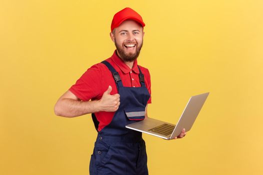 Satisfied worker man wearing blue overalls, red T-shirt and cap working on laptop, showing thumb up, likes a new online service, looking at camera. Indoor studio shot isolated on yellow background.