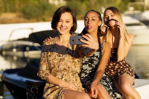 Smiling ethnic woman taking self portrait on cellphone with black girlfriend showing tongue and blonde making grimace on pier