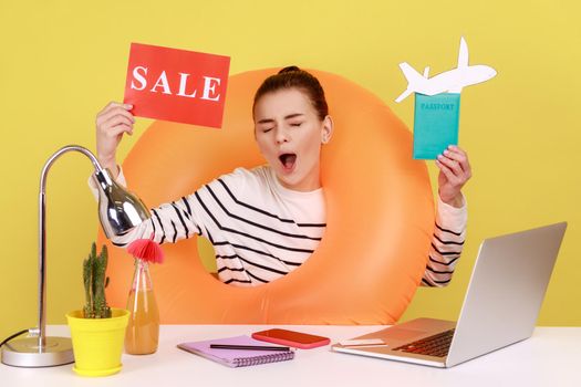 Joyful woman with rubber ring, holding card with sale inscription, passport and paper plane, rejoice sales for summer tours. Indoor studio studio shot isolated on yellow background.