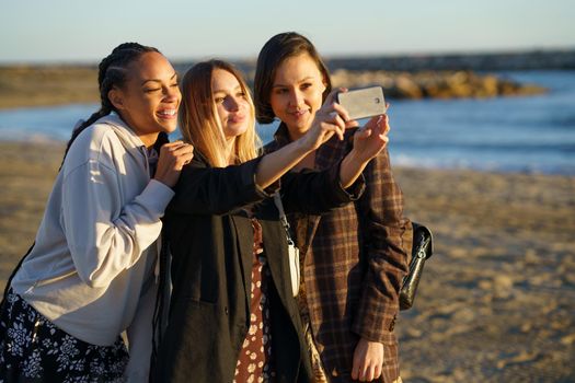 Positive multiethnic female friends taking self portrait on modern smartphone while standing on sandy shore near sea on sunny day