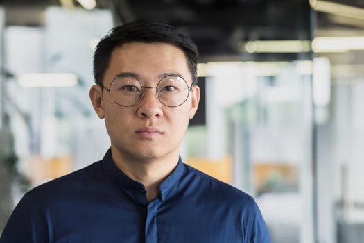 Closeup portrait of successful asian investor in glasses and blue shirt, businessman looking thoughtfully at camera, man working inside modern office building, experienced and mature boss manager.