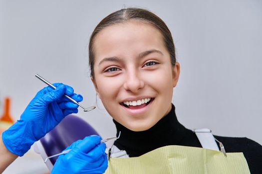 Portrait of young teenage girl in dental chair with hands of doctor with tools. Female teenager smiling with teeth looking at camera in dentist office. Adolescence hygiene treatment dental health care