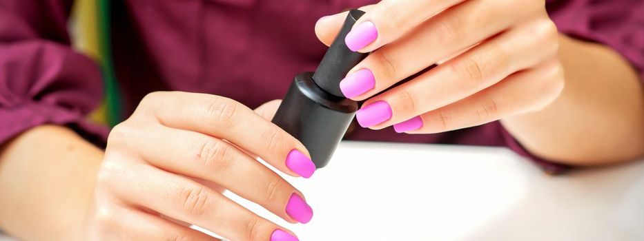 White woman holding nail polish black bottle with painted pink nails close up