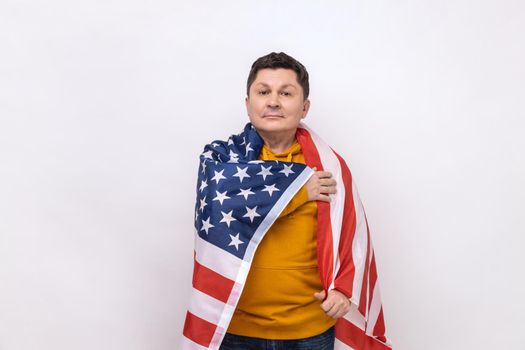Serious man being wrapped in USA flag, celebrating labor day or US Independence day 4th of july, holding hand on chest, wearing urban style hoodie. Indoor studio shot isolated on white background.