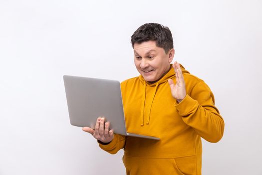 Man waving hand gesturing hello to laptop screen, talking on video call with coworker, communicating online conference, wearing urban style hoodie. Indoor studio shot isolated on white background.