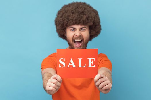 Portrait of excited crazy man with Afro hairstyle wearing orange T-shirt standing showing sale card, presenting low prices, shopping. Indoor studio shot isolated on blue background.