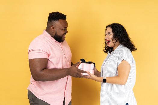 Portrait of romantic young couple in casual clothing standing together, husband giving present box, congratulating with anniversary. Indoor studio shot isolated on yellow background.