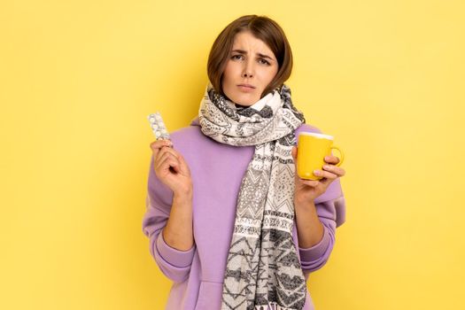 Portrait of sick woman wrapped in scarf holding pills and tea cup, screaming, feeling annoyed from virus grippe, wearing purple hoodie. Indoor studio shot isolated on yellow background.