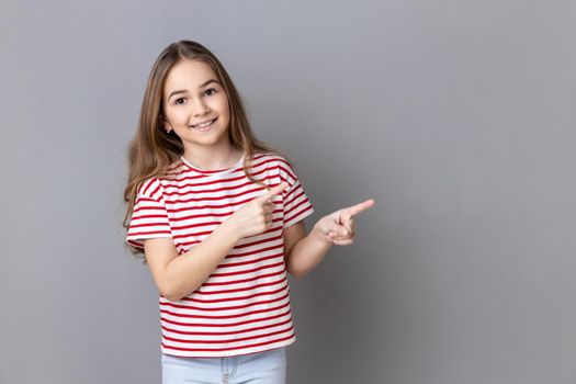 Attention to advertising. Little girl wearing striped T-shirt pointing aside, showing copy space for commercial text, blank wall with idea presentation. Indoor studio shot isolated on gray background.