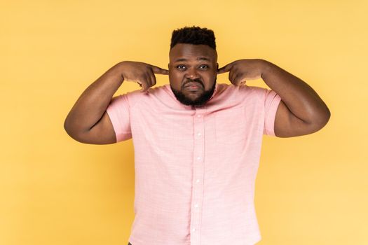 I dont want to listen it. Annoyed man hears loud noise, avoids unpleasant sound, doesnt hear somebody's advice, closes ears with index fingers. Indoor studio shot isolated on yellow background.
