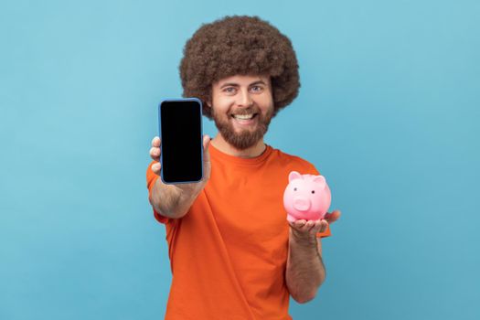 Portrait of happy satisfied man with Afro hairstyle in orange T-shirt holding in hands piggy bank and smart phone with empty screen for advertisement. Indoor studio shot isolated on blue background.