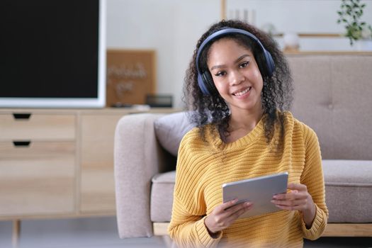 Young satisfied female in casual clothes yellow and headphones surfing digital tablet while sitting on floor with legs crossed near soft couch