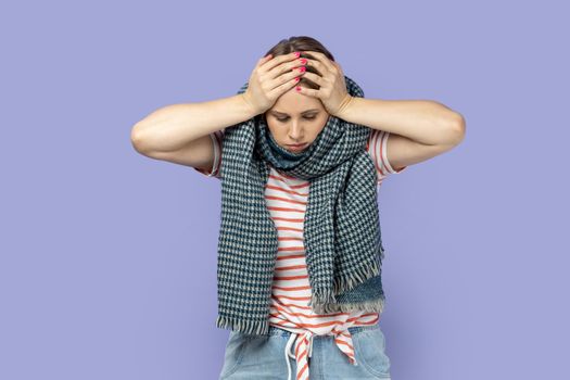 Portrait of sick unhealthy woman wearing striped T-shirt feels unwell, suffering terrible headache, standing wrapped in warm scarf, having influenza. Indoor studio shot isolated on purple background.