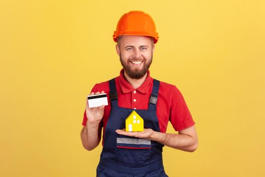 Portrait of happy builder man wearing blue uniform and protective helmet showing credit card and paper house, looking at camera with smile. Indoor studio shot isolated on yellow background.