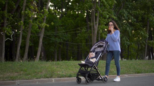 Mom walks with a baby in a stroller and eats a burger on the go. A young woman in glasses pushes a stroller with her son and eats a burger in the park. 4k